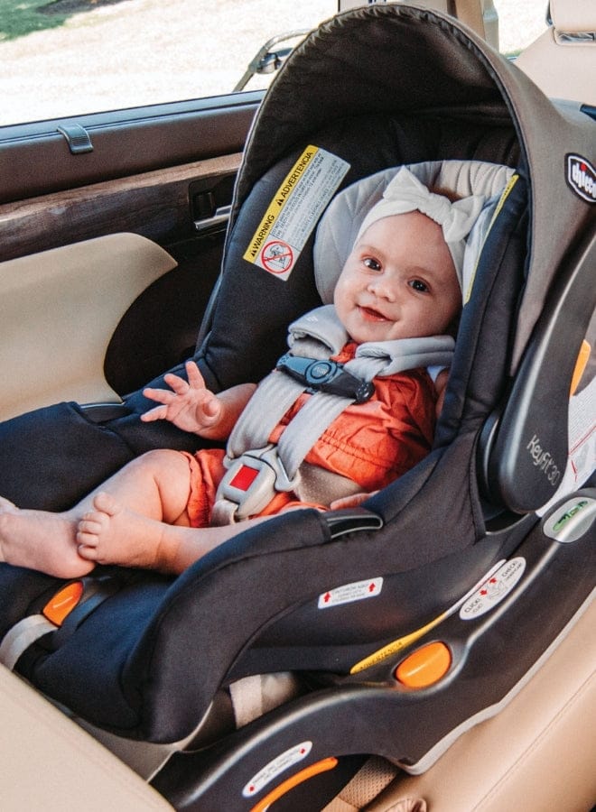 A baby wearing a white headband sits smiling in a black Chicco car seat with gray straps, buckled in a car. A yellow warning sticker is visible.