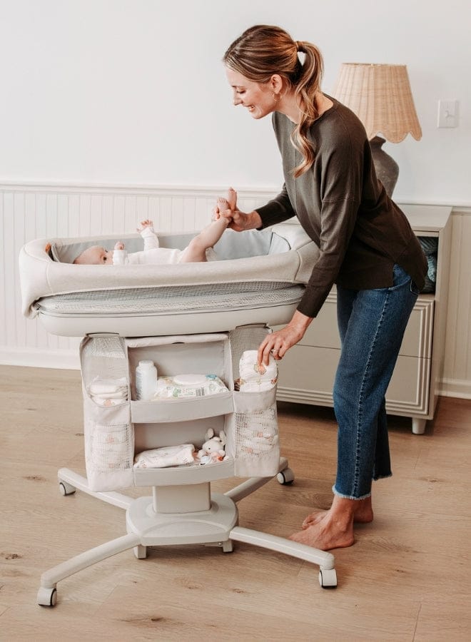 A woman stands next to a baby in a bedside bassinet, smiling and holding the baby's leg. The bassinet has storage compartments.
