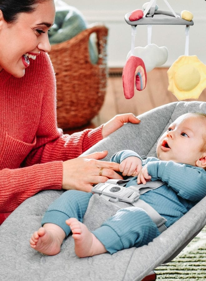 A woman smiles at a baby lying in a gray bouncer under a toy mobile with a rainbow, sun, and cloud. The woman wears a red sweater.