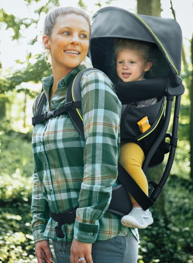 A smiling woman wearing a green plaid shirt carries a toddler in a gray backpack carrier through a sun-dappled forest.
