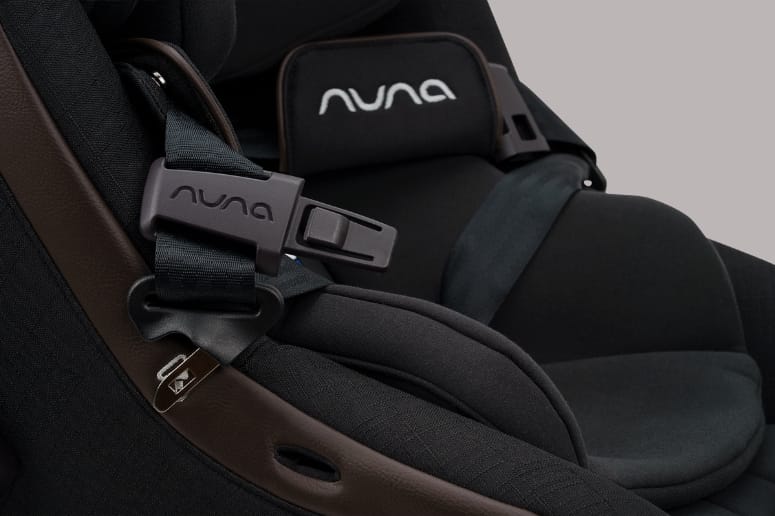 Close-up of black Nuna car seat with brown trim, showing harness buckle and brand logo on headrest and buckle.