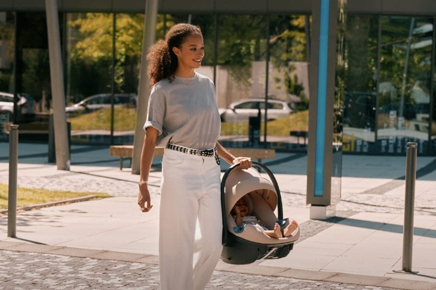 A woman carries a baby in a car seat in a sunny outdoor urban setting. She wears casual clothes and smiles.