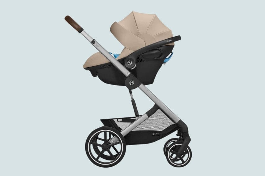 A tan Cybex infant car seat sits in a gray stroller on a blue background. The stroller has black wheels, a brown handle, and a black storage basket.