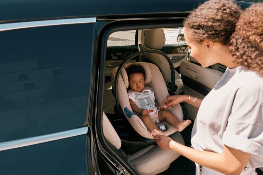 A mother secures her baby in a car seat inside a dark car. The baby is in the back seat. Natural lighting.