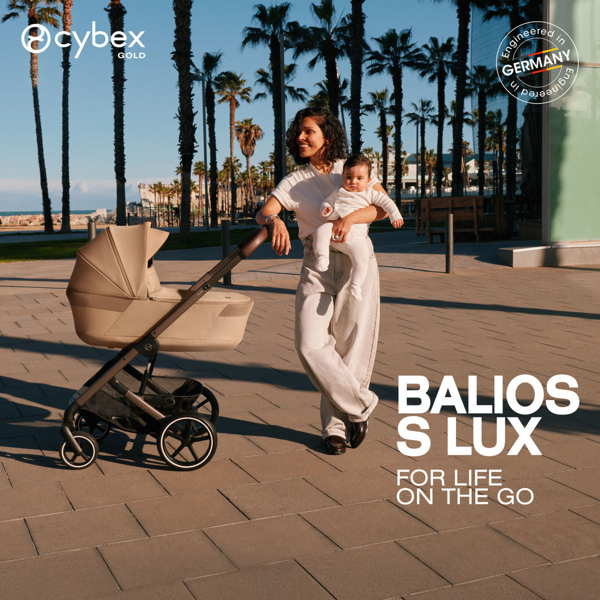 Woman holding a baby next to a beige Cybex Balios S Lux stroller on a palm tree lined path