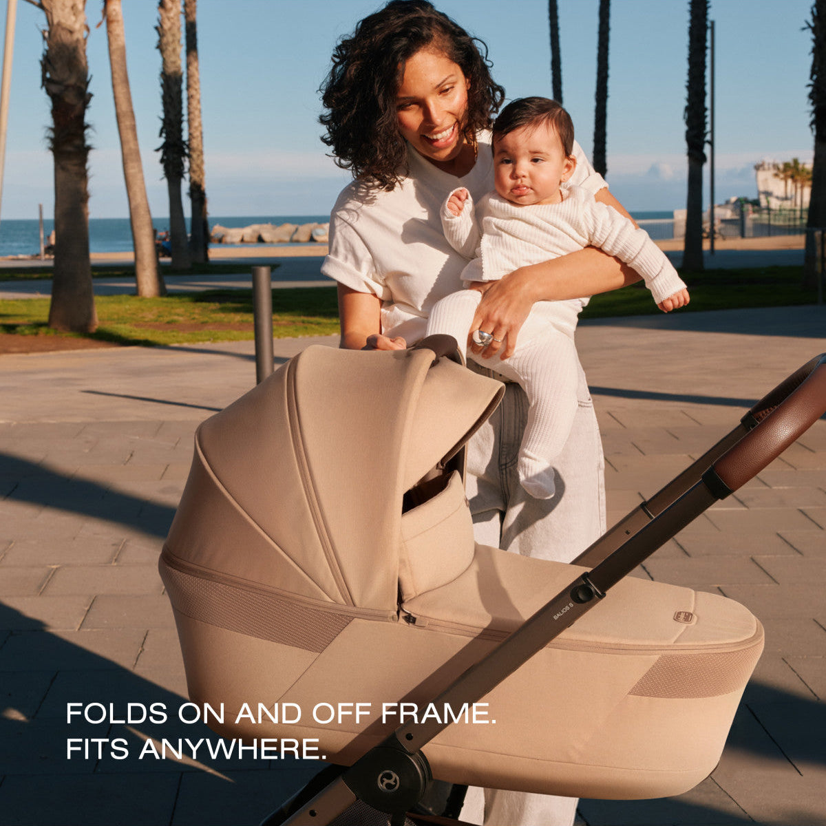Woman holding baby next to a beige Cybex Balios S stroller carrycot on a paved path