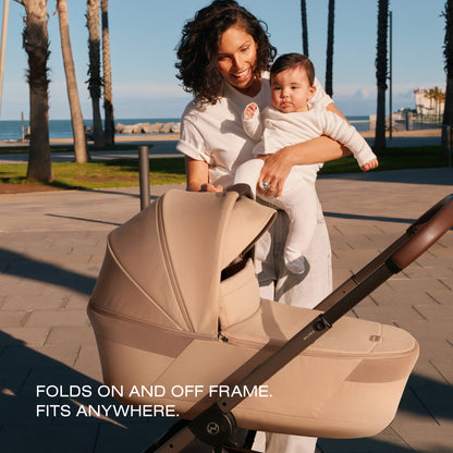 Woman holding baby next to a beige Cybex Balios S stroller carrycot on a paved path