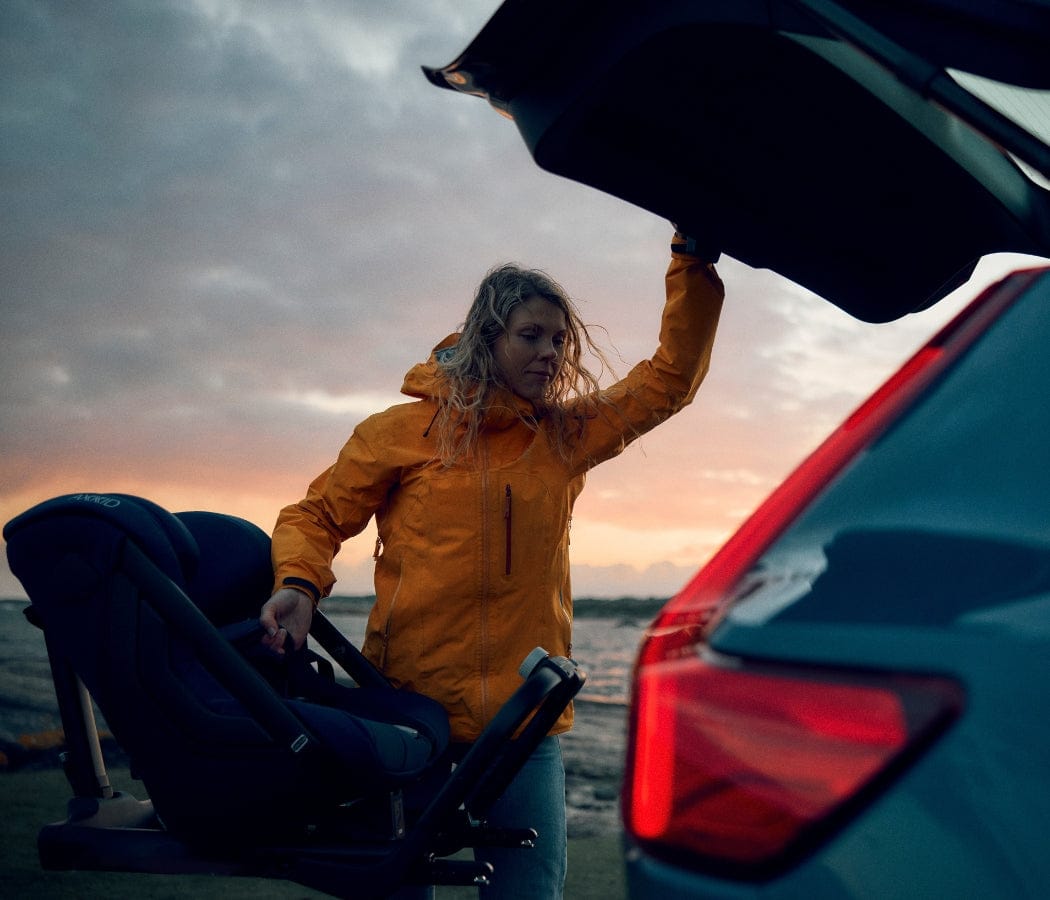Woman in orange jacket loads black car seat into open trunk of blue car at sunset.