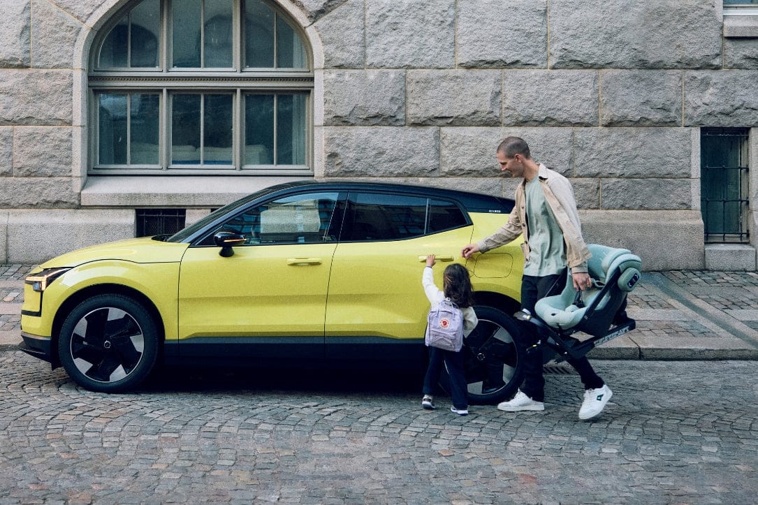 Bright yellow electric car parked on cobblestone street; man carries child seat, child touches car door, stone building in background.