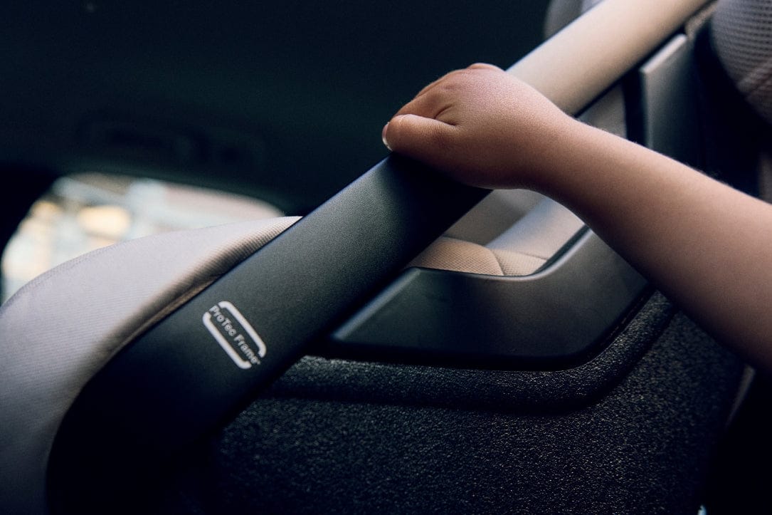 Child’s hand gripping black car seat handle with “ProTec Frame” logo, beige seat, dim interior lighting.