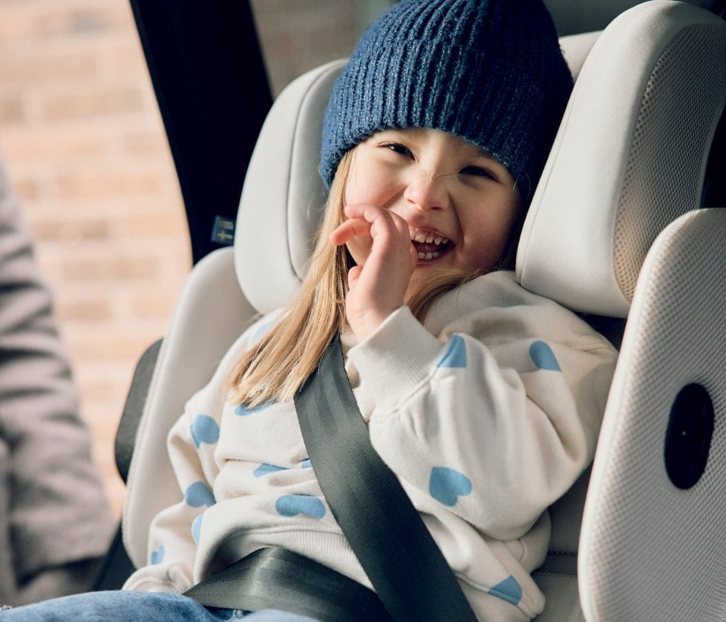 Smiling toddler in blue beanie and heart-patterned sweater, secured in white car seat, hand near mouth, bright natural light.