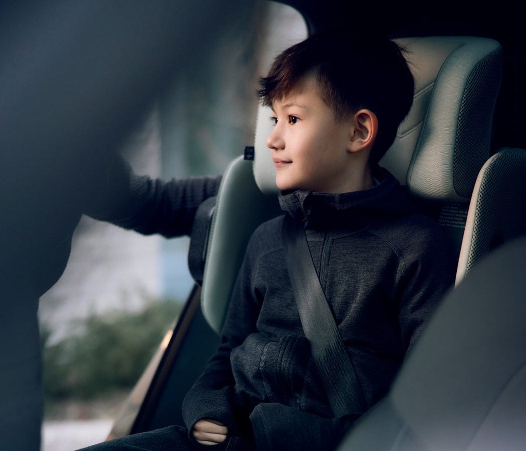 Boy in gray hoodie sits in car seat, gazing out window with soft smile, hand resting on door.