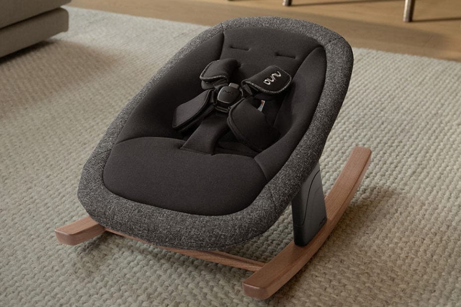 Dark gray fabric baby rocker with wooden base, black harness, and "Nuna" logo, on beige textured rug.