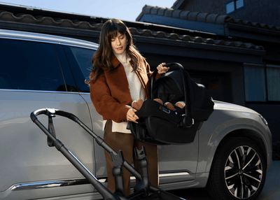 Woman in brown coat attaches black infant car seat to stroller beside silver SUV, sunny day.