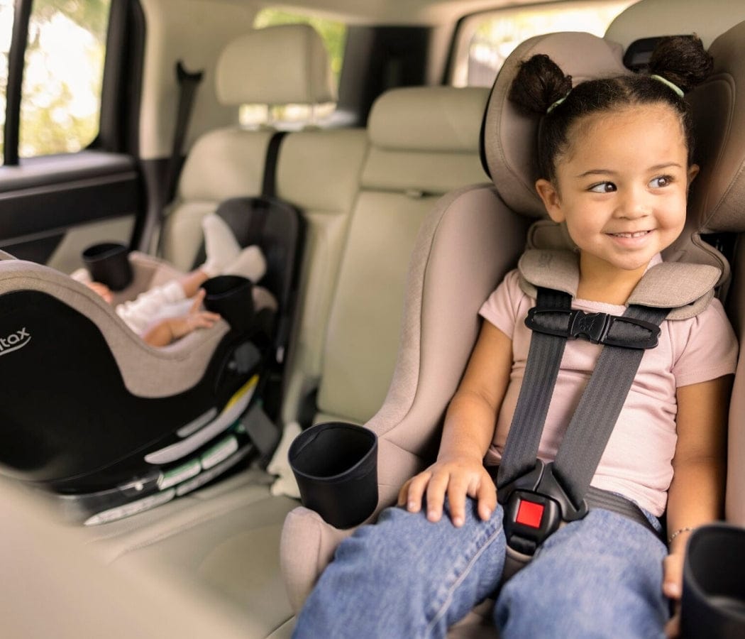 A smiling child sits in a car seat, buckled in. A baby in a car seat is in the background. The interior is light-colored.