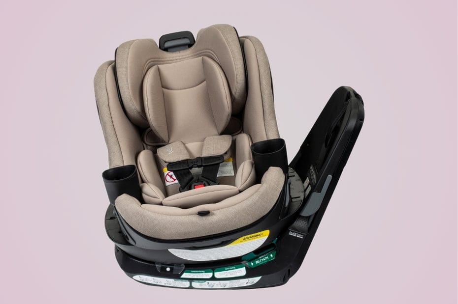 Tan and black car seat with safety straps and two cup holders, angled on a pink background. Text is visible on the bottom portion.