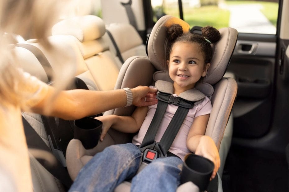 A smiling young child sits securely in a car seat, with an adult adjusting the harness. The seat is gray, complementing the car's beige interior.