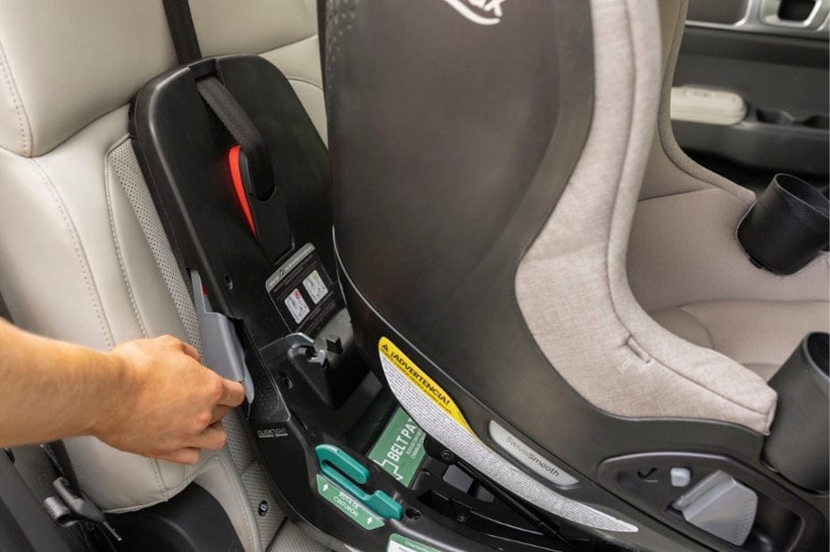 A person's hand pushes a gray latch on a black car seat base with the attached car seat. The seat is light gray, and the seats in the car are light gray.