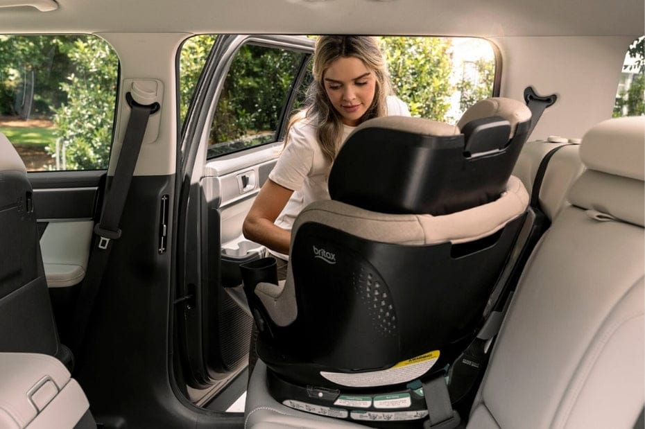 A woman installs a black and tan Britax car seat in the backseat of a car, surrounded by light gray leather interior. Green trees are visible through the car window.