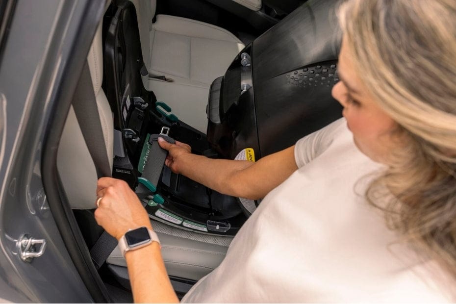 A woman installs a car seat in the back of a grey car, securing it with a seatbelt. The car seat is black and grey. The seat is white leather.