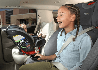 Smiling girl in gray Britax car seat, wearing denim shirt, secured with seatbelt, holding cup; another child in rear-facing seat behind.
