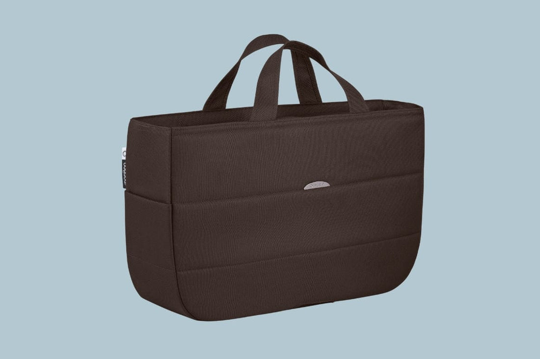 Brown textured tote bag with dual handles, front pocket, and silver logo tag, set against light blue background.