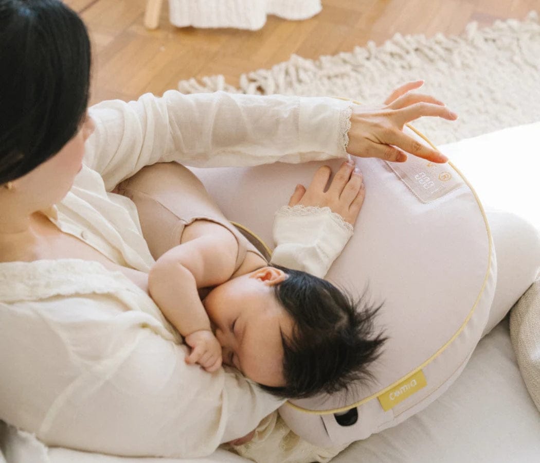 A baby nurses on a light-skinned person's lap, supported by a pink and yellow nursing pillow. The person's hand gently rests on the pillow. "Comfy" is printed on it.