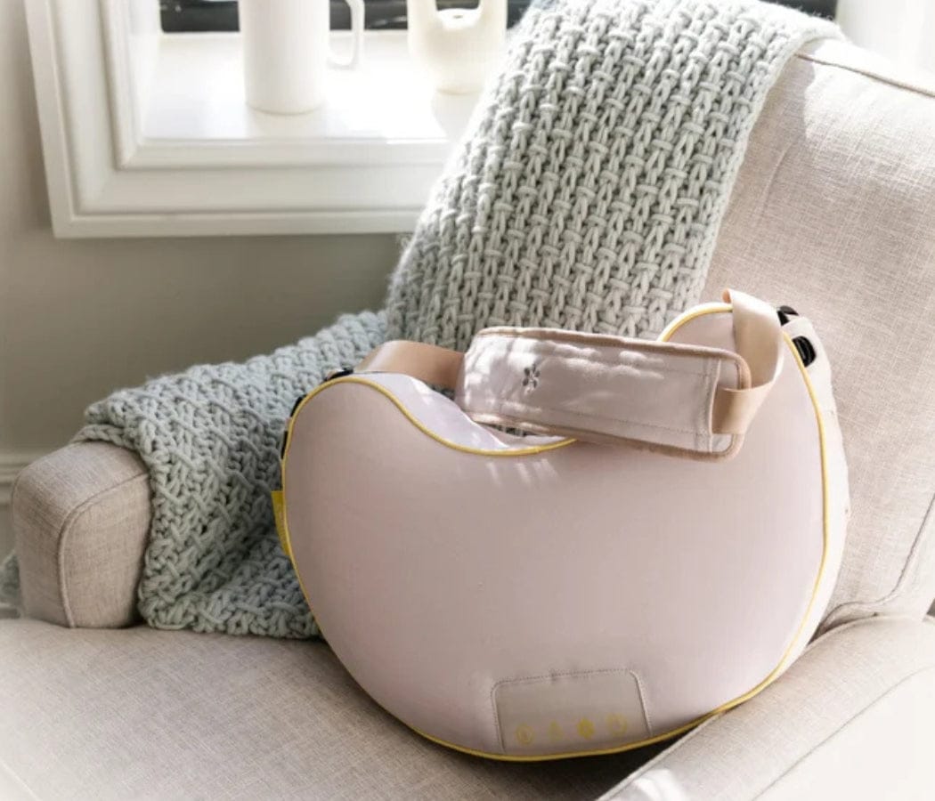 A light pink massage pillow with a yellow trim sits on a beige armchair with a light gray throw blanket.