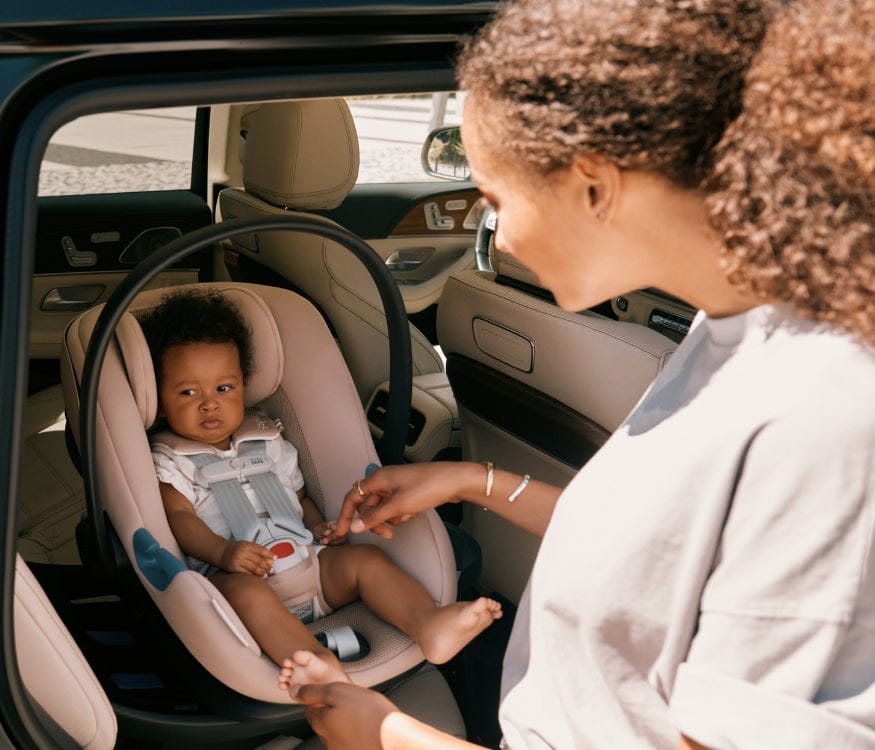 A parent buckles their baby into a light pink car seat in a tan-colored car. The baby looks forward, and the parent's hands are visible.