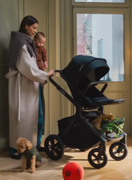 Woman holding baby stands beside black stroller with groceries, small dog nearby, red ball on floor, warm indoor lighting.