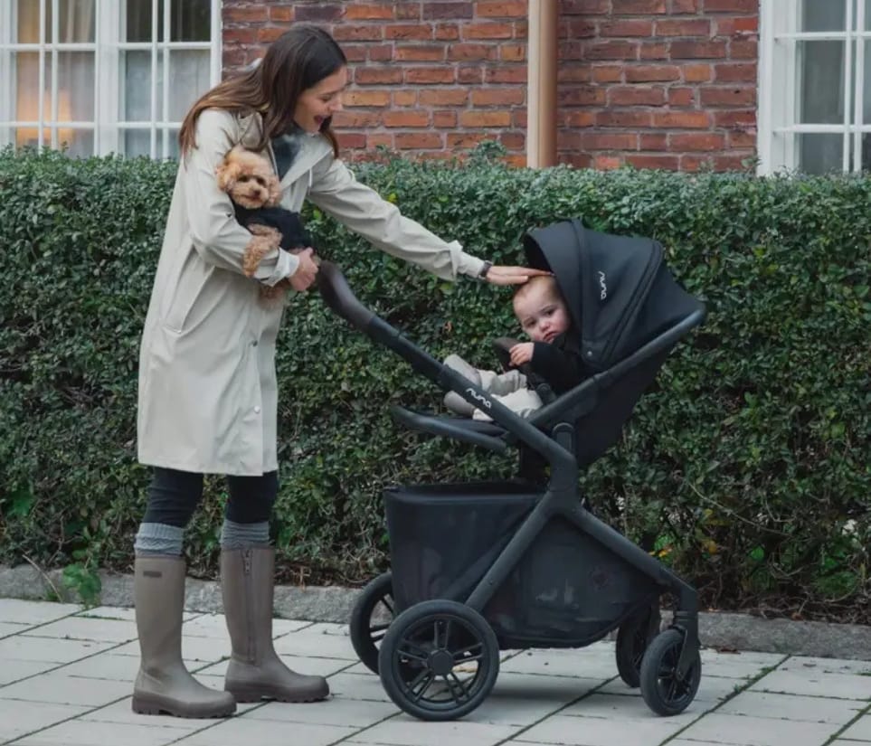 Woman in beige coat holds small dog while pushing black stroller with baby, standing on paved path near green hedge and brick building.