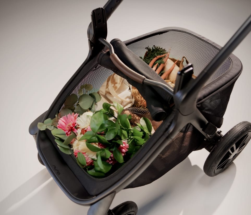 Black stroller basket filled with pink flowers, green leaves, carrots, and fabric, viewed from above on a white background.