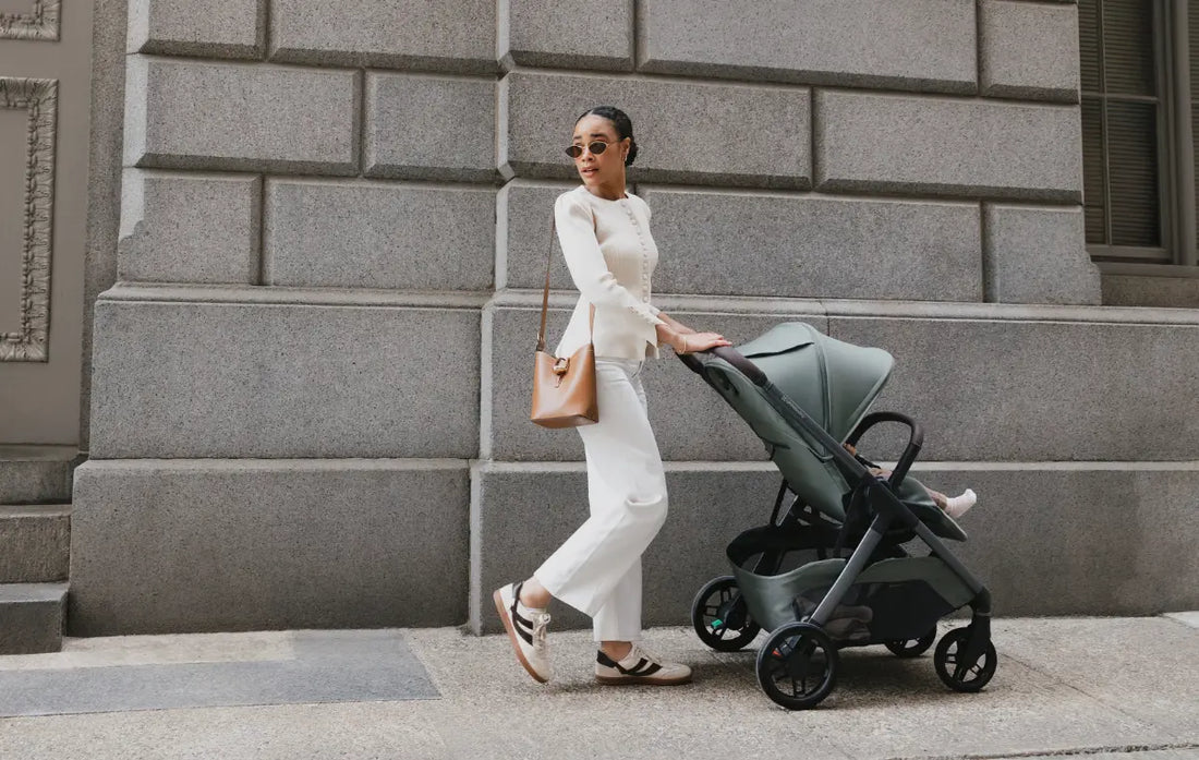 woman standing with kona stroller looking back against a beige block wall. new- Kona from UPPAbaby now for pre-order at Albee Baby!