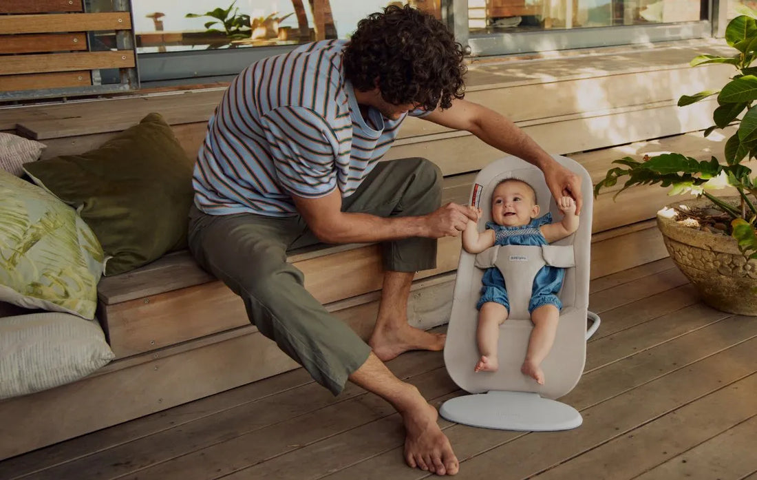 dad sitting next to baby in babybjorn bouncer begin outside on a deck - new at albee!