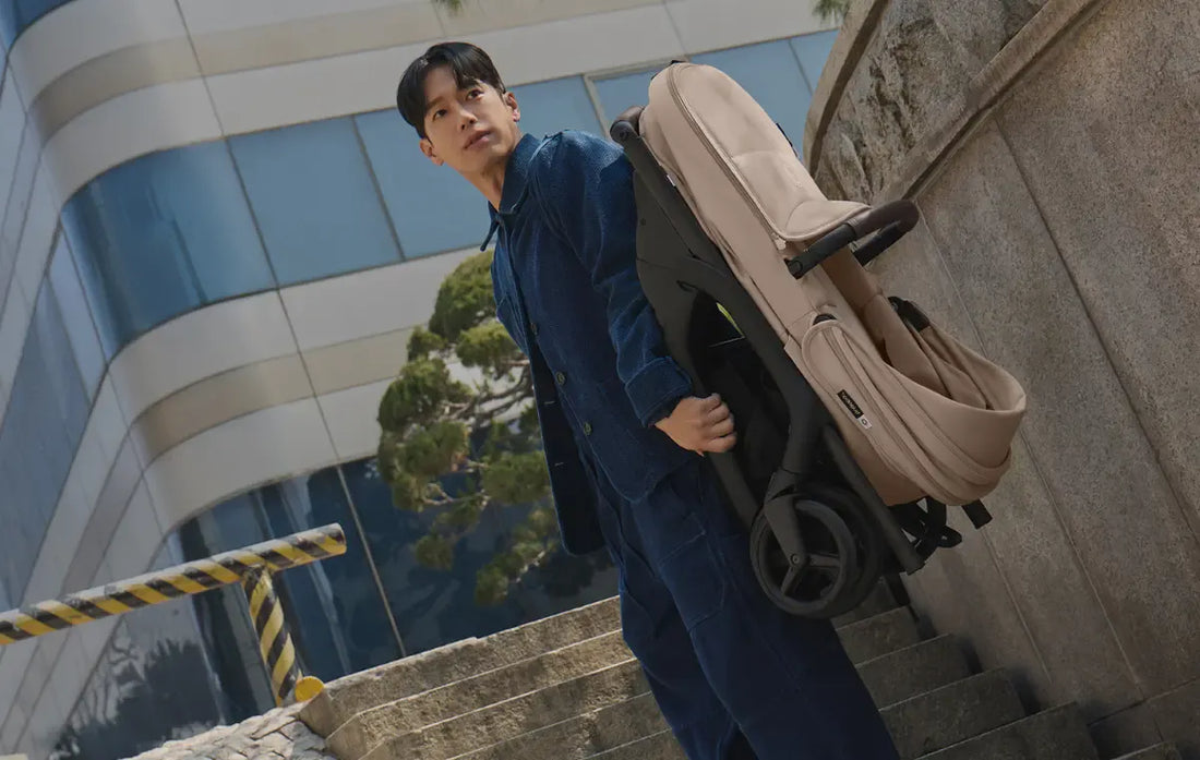 A man in a navy blue jacket and matching pants carries a beige and black baby stroller on his shoulder while climbing outdoor stone steps, with a modern glass building and yellow-and-black striped barrier visible in the background.