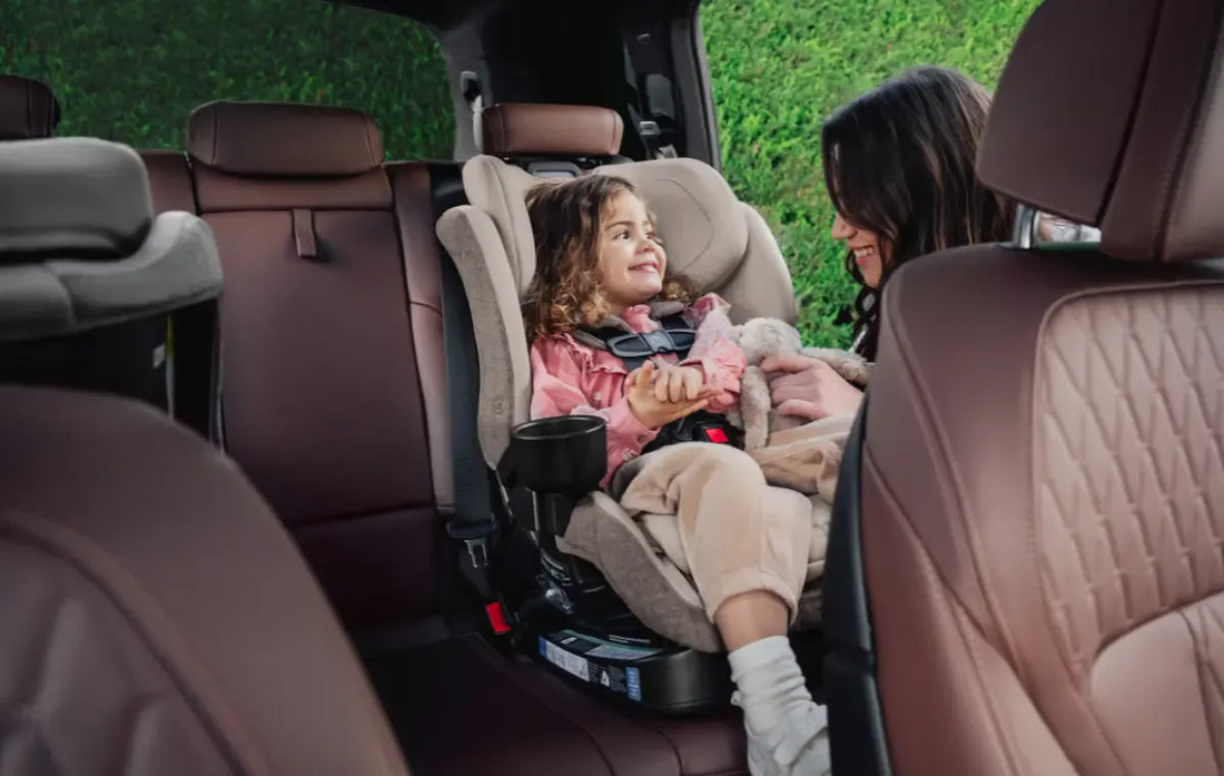 Mother smiling and interacting with a young child secured in a forward-facing car seat inside a vehicle. The child holds a stuffed toy and looks happily toward the parent, with spacious brown leather seating visible throughout the car interior.