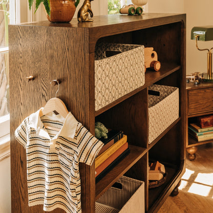 Babyletto dark wood bookshelf with storage bins and a striped shirt hanging on the side hook