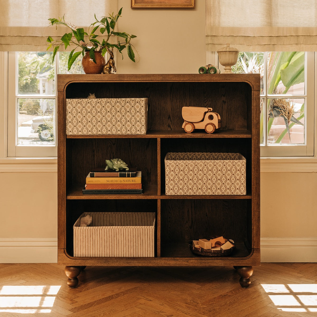 Babyletto dark wood bookshelf with four cubbies containing patterned storage bins and wooden toys