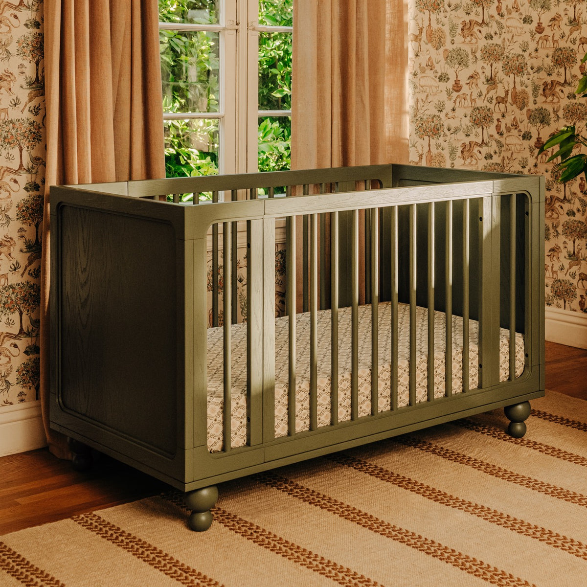 Babyletto olive green wooden crib with spindle sides on a patterned rug in a nursery