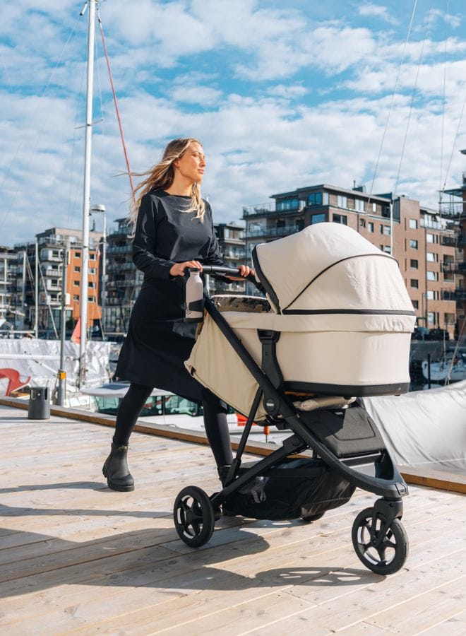 Woman in black dress pushes cream baby stroller on wooden dock, modern buildings and boats in background under blue sky.