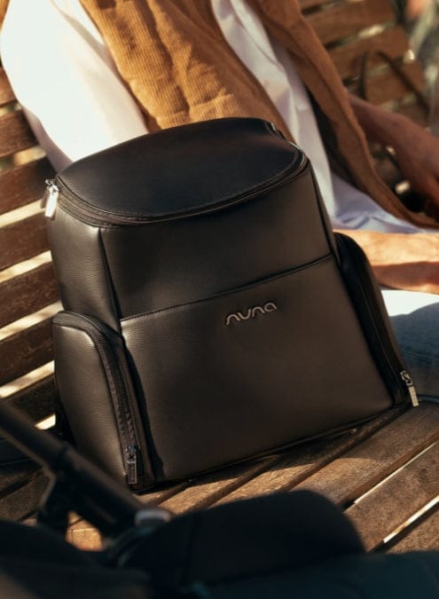 A black Nuna diaper backpack sits on a wooden park bench next to a baby stroller. A person in a brown sweater sits behind.