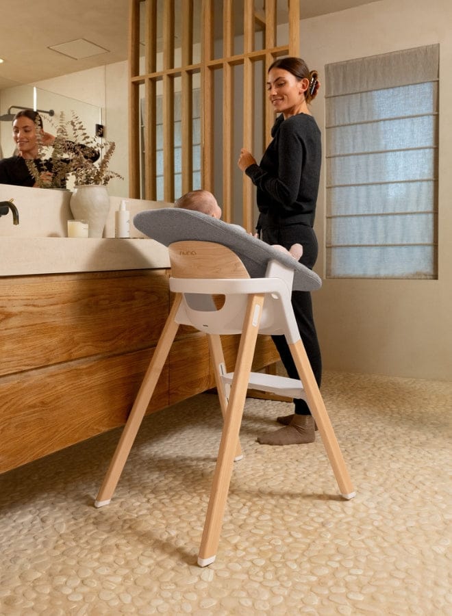 Woman stands beside baby in modern wooden high chair with gray cushion, in bright, minimalist bathroom with stone floor and wooden vanity.