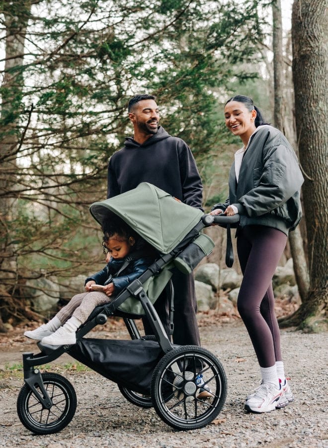 Family of three with child in stroller walking in wooded park, smiling, natural light, green canopy, gravel path.
