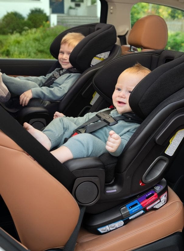 Two children sit in black car seats on brown leather seats. A house and trees are in the background. The children wear blue outfits.