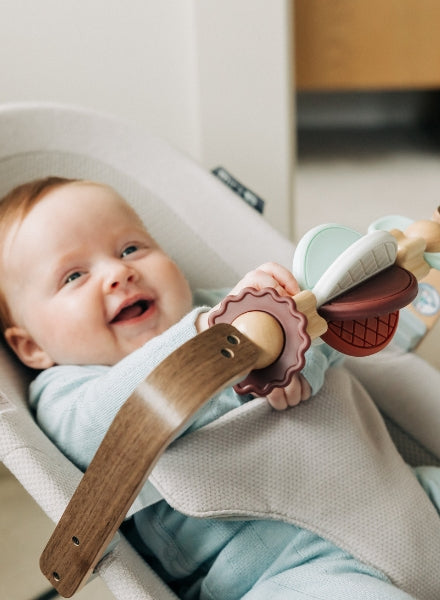 Baby sitting in a bouncer seat playing with a wooden and silicone toy bar