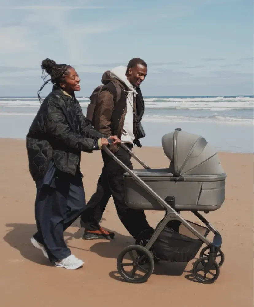 man and woman pushing silver cross cove 2 stroller with bassinet on a beach.