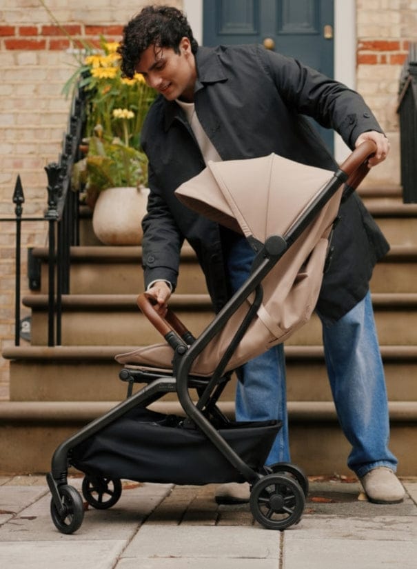 A man on stone steps adjusts a brown baby stroller with a black basket underneath. He wears a dark coat and jeans.