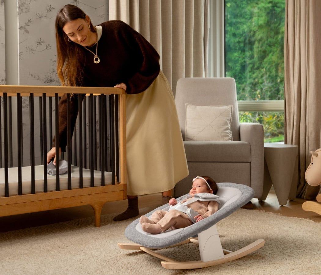 Woman tending to baby in crib, infant resting in gray rocker, cozy nursery with natural light and neutral tones.