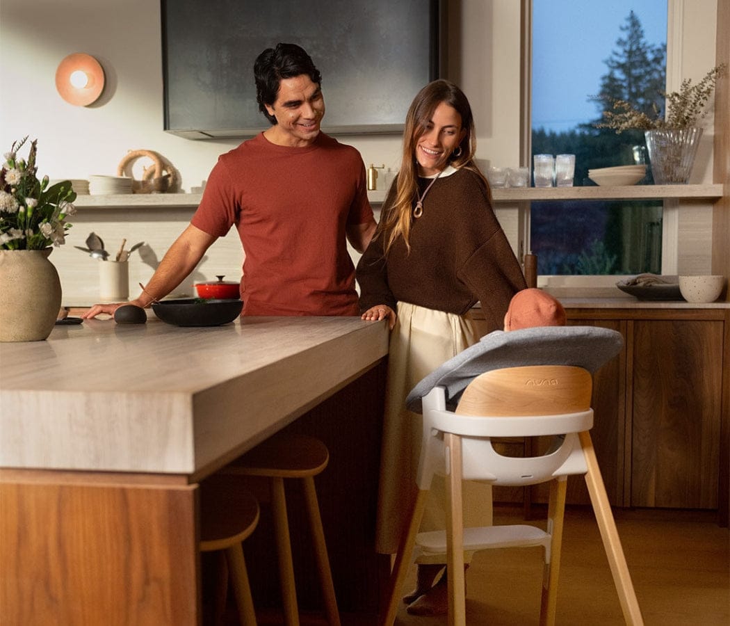 Smiling couple in warm-toned kitchen with wooden highchair in foreground, soft lighting, window view of trees.