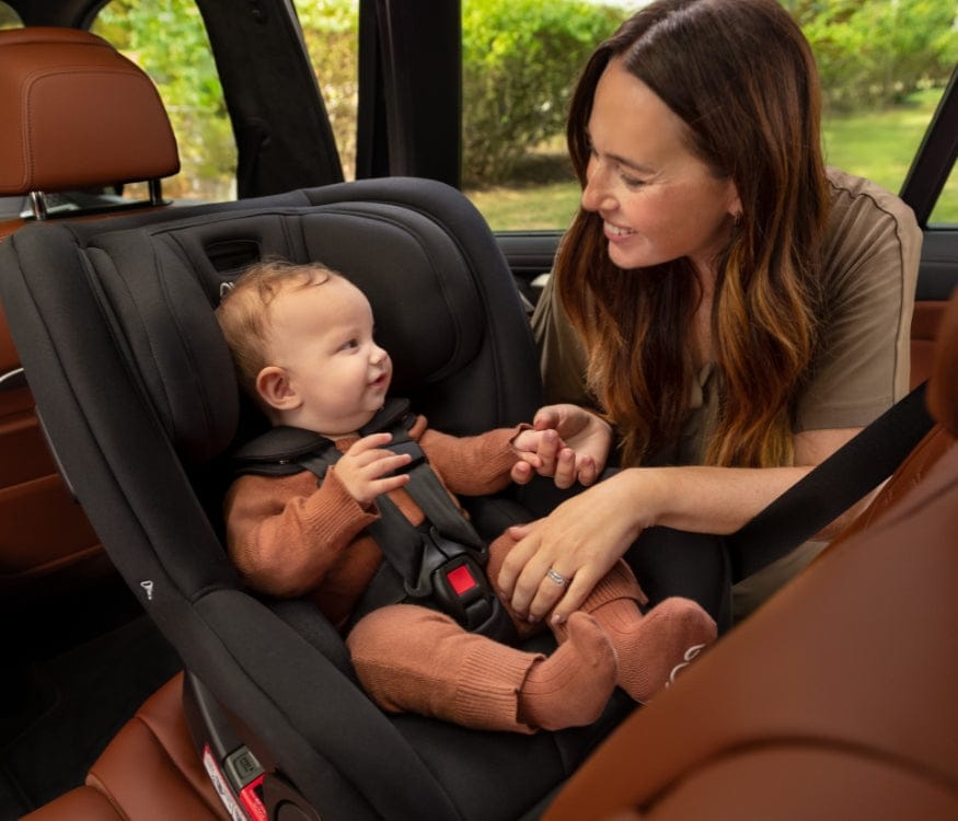 A baby sits in a black car seat, secured with a safety harness, while a smiling woman holds the baby's hand.